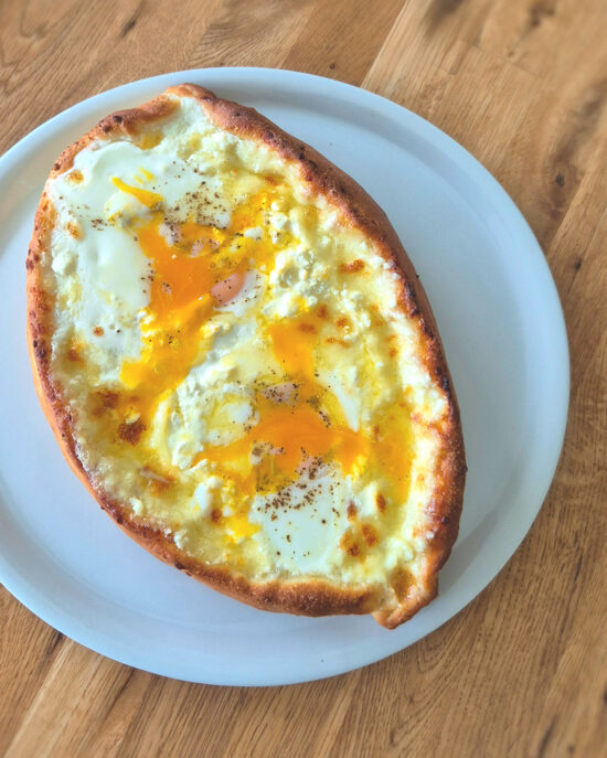 Adscharuli Khachapuri in Bootsform mit goldbraunem Teigrand, cremigem Käse und flüssigem Eigelb auf einem weissen Teller.