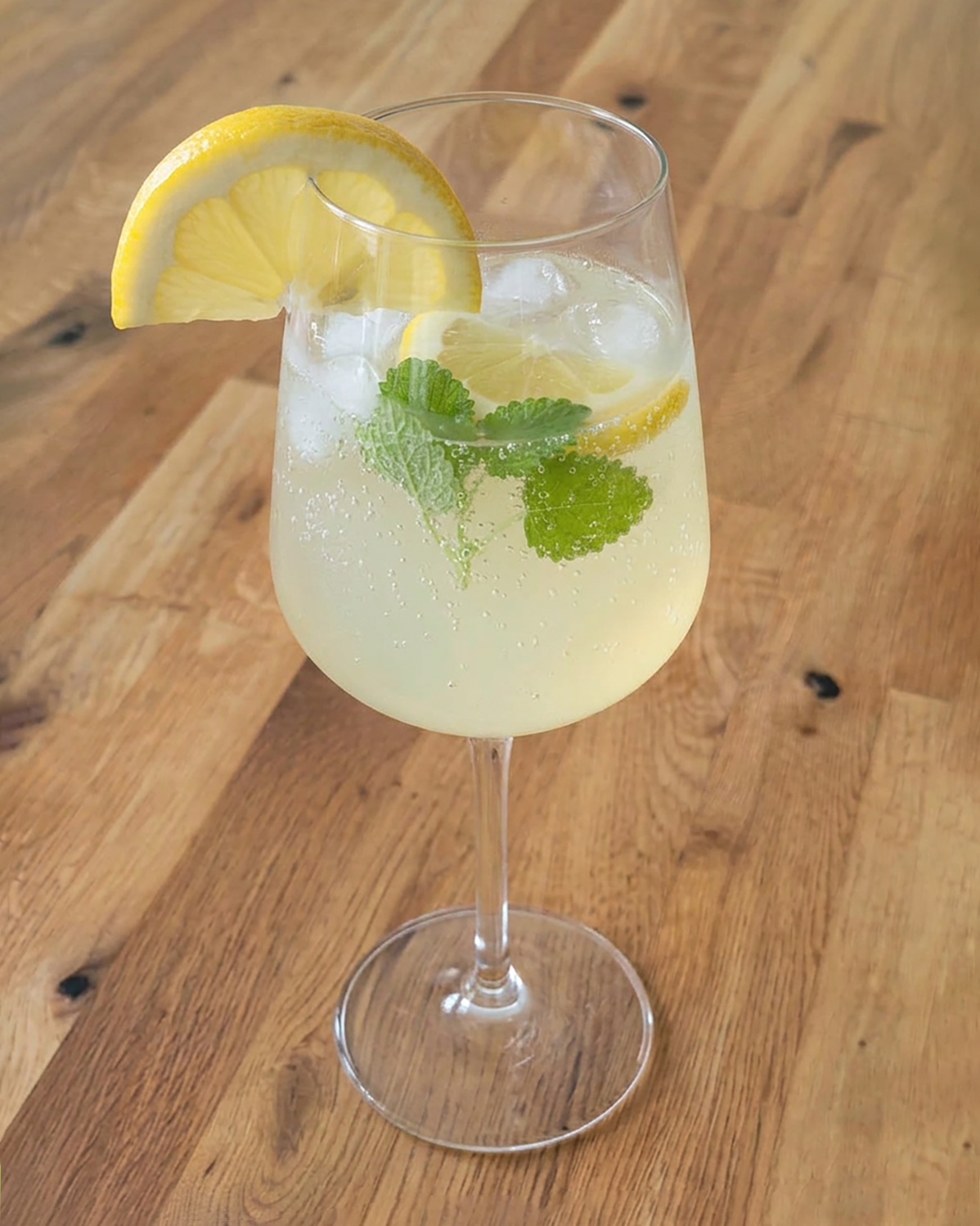 Ein zitroniger Limoncello Spritz mit Prosecco, Eiswürfeln, Zitronenscheibe und Melisse im eleganten Glas auf Holztisch