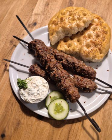 drei Adana Kebap Spiesse mit halbiertem Fladenbrot, einer Schale Cacik, Petersilie und Gurken
