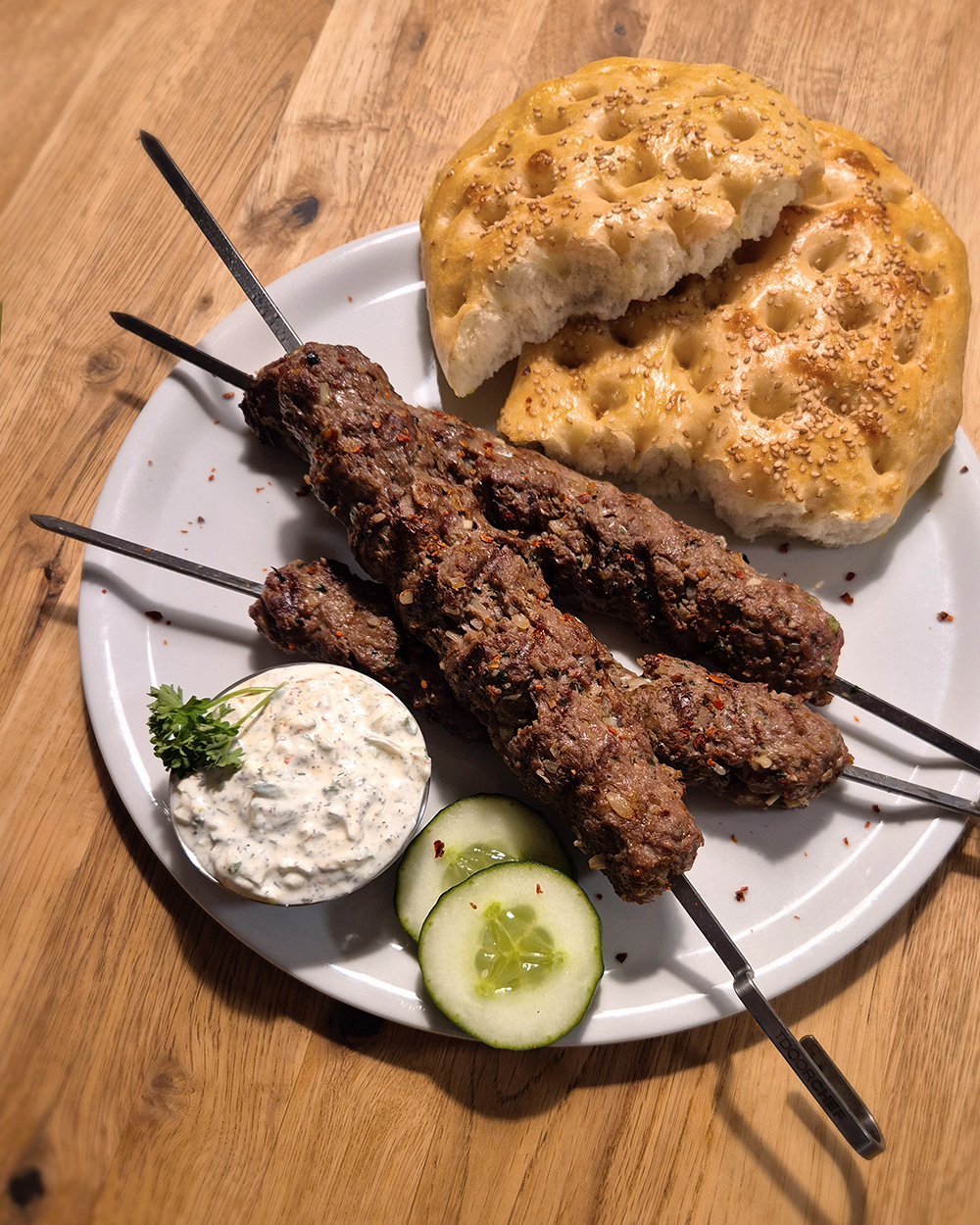 drei Adana Kebap Spiesse mit halbiertem Fladenbrot, einer Schale Cacik, Petersilie und Gurken