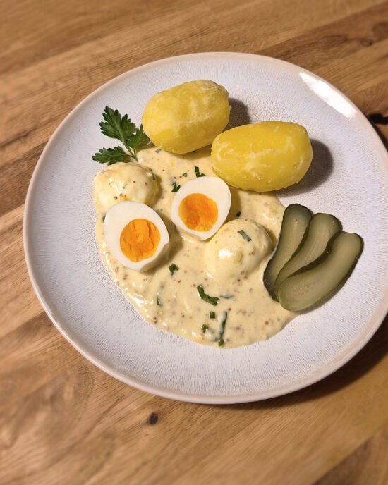 Gekochte Eier in Senfsauce serviert mit Salzkartoffeln und Gewürzgurken auf einem Teller.