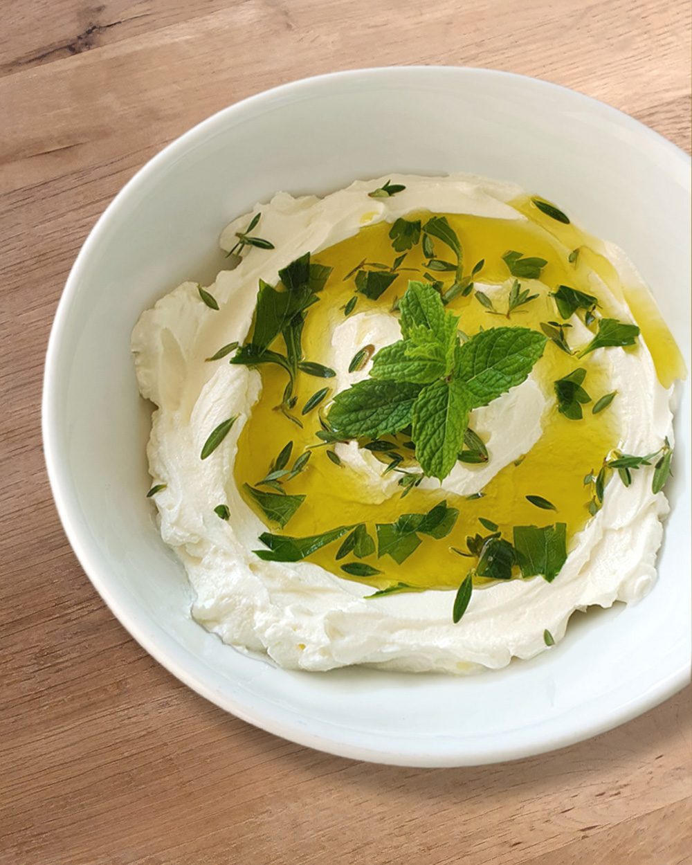 Schüssel mit Labneh, garniert mit Olivenöl, frischen Kräutern und Minze auf einem Holzuntergrund.