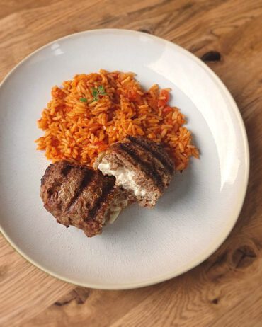 Bifteki auf einem Teller, serviert mit einer Portion Tomatenreis. Das Bifteki ist gegrillt und aufgeschnitten.