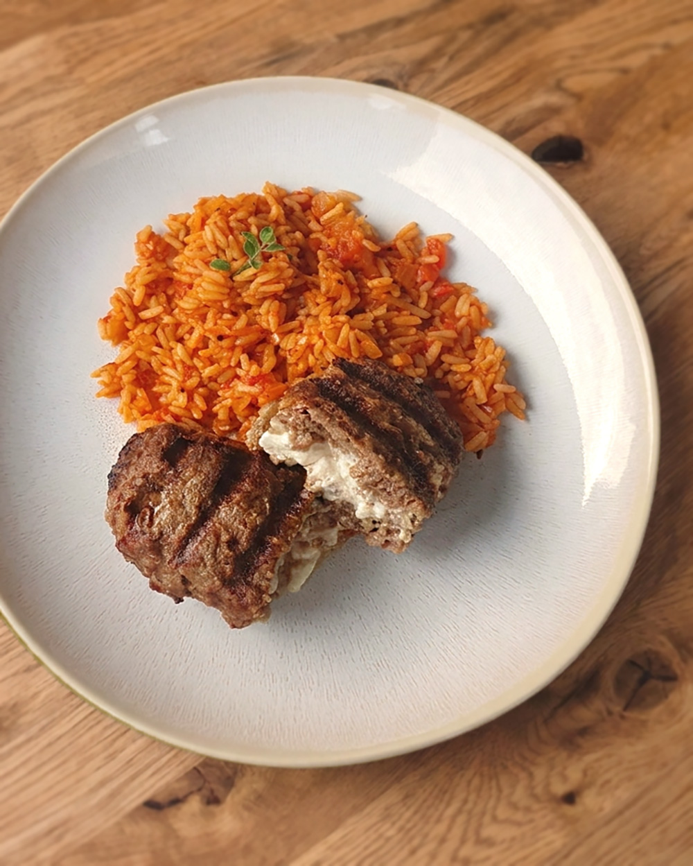 Bifteki auf einem Teller, serviert mit einer Portion Tomatenreis. Das Bifteki ist gegrillt und aufgeschnitten.