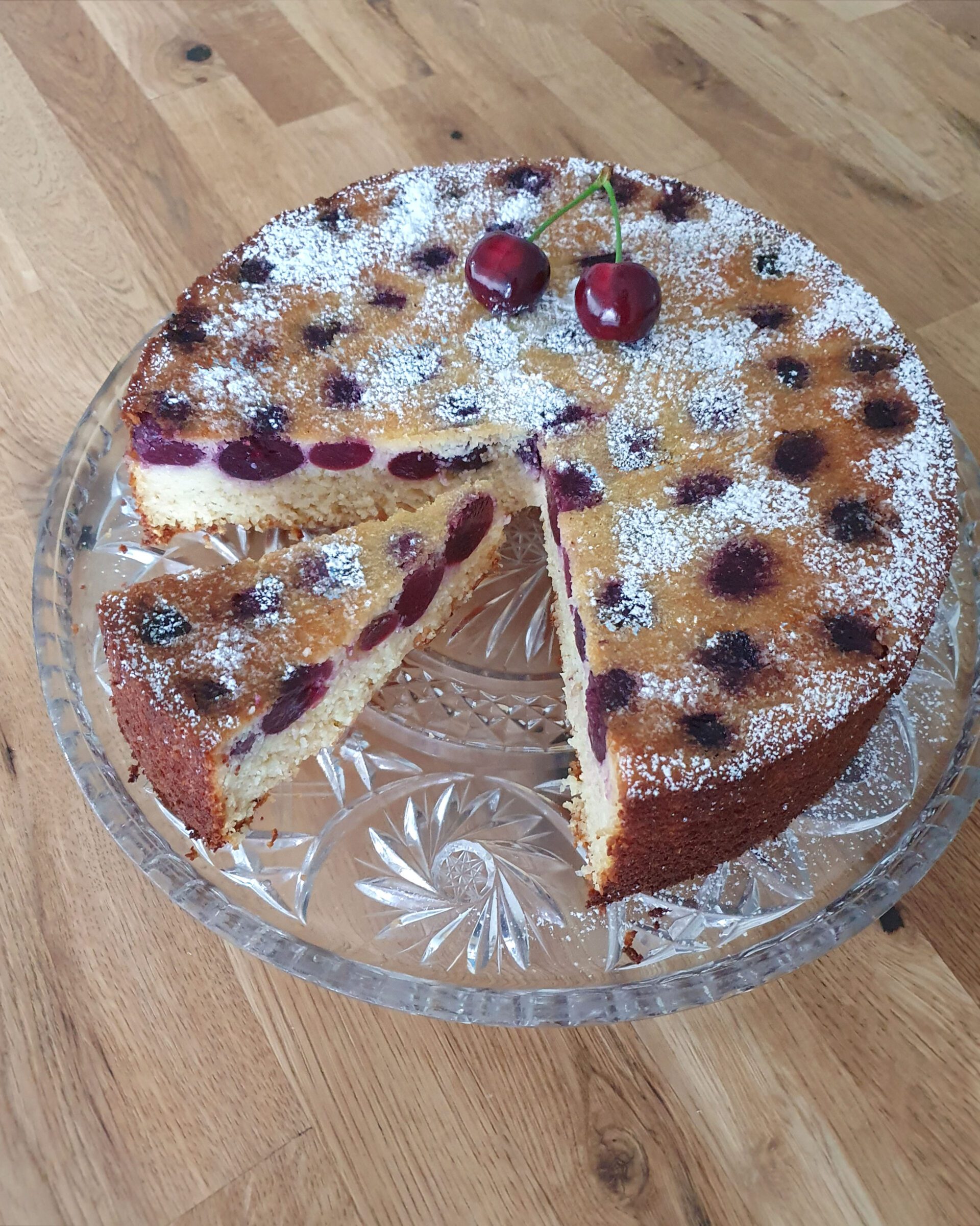 Saftiger Griesskuchen mit frischen Kirschen und Haselnuss, mit Puderzucker bestäubt, ein Stück angeschnitten, auf einem Glasteller