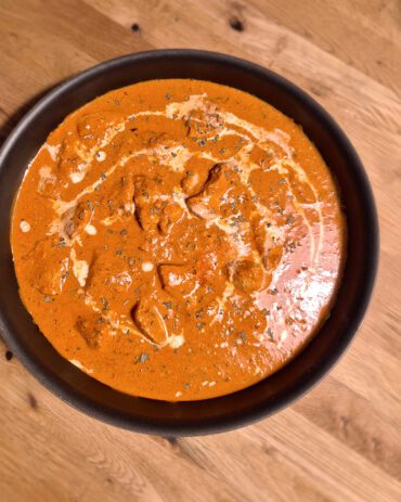 Butter Chicken mit Sahnestrudel und Bockshornkleeblätter garniert in einer schwarzen Pfanne auf Holzuntergrund