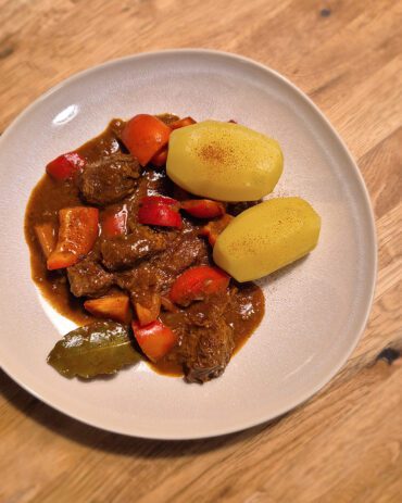 Gulasch mit Paprika und Salzkartoffeln auf einem Teller, garniert mit einem Lorbeerblatt.