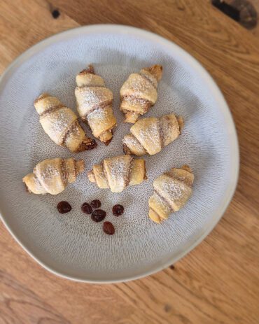 Walnuss-Rugelach auf einem Teller, mit Puderzucker bestäubt, umgeben von einigen Rosinen.