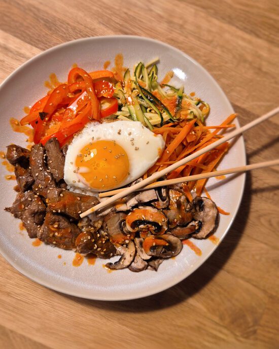 Schüssel mit Bibimbap, bestehend aus Rindfleisch, Spiegelei, Karotten, Zucchini, Paprika und Champignons, garniert mit Sesam.