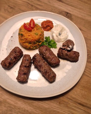 Cevapcici auf einem Teller, serviert mit einer Portion Djuvec Reis, Tomatensauce und Joghurt-Dip, garniert mit Kirschtomaten und frischer Petersilie.