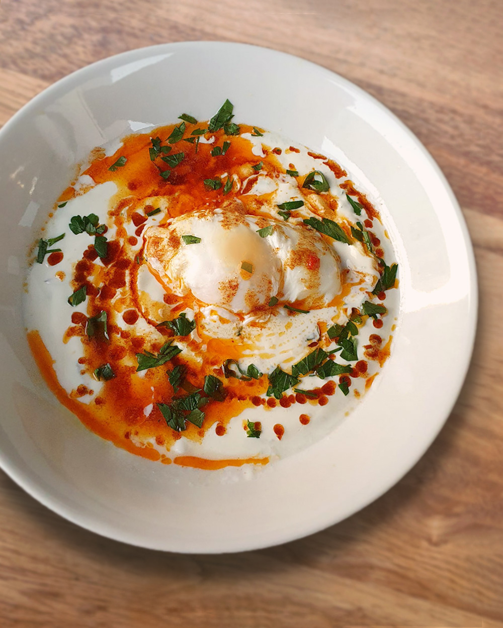 Cilbir, ein Gericht mit pochierten Eiern auf Joghurt, garniert mit Paprikabutter und frischen Kräutern in einer weißen Schüssel.