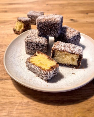 Sechs quadratische Kuchenstücke mit einem hellen Biskuit, umhüllt von Schokolade und Kokosraspeln, auf einem Teller.