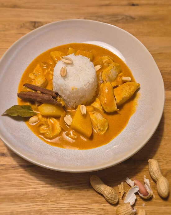 Hähnchen Massaman-Curry mit Reis, Erdnüssen und Gewürzen auf einem Teller, serviert auf einem Holzuntergrund.