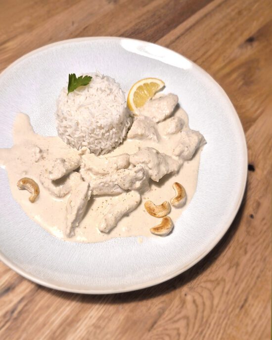 Malai-Chicken serviert mit Reis, einer Zitronenscheibe und Cashewkernen auf einem weißen Teller.