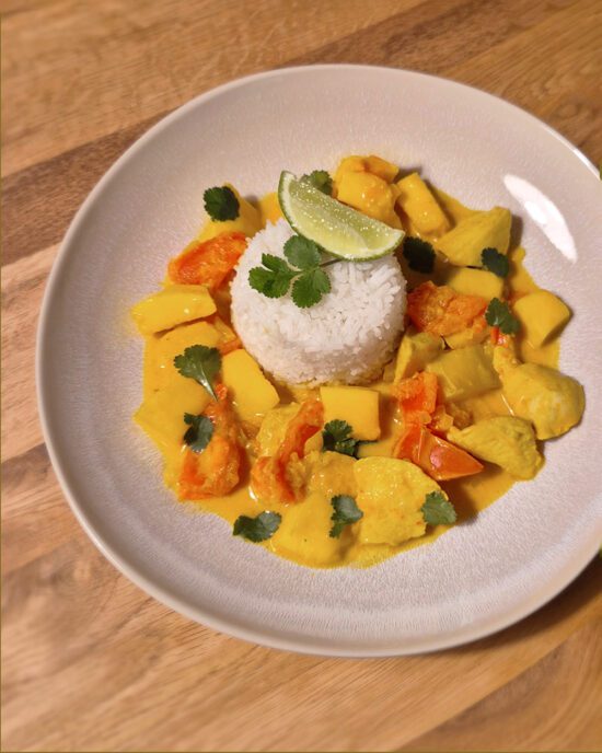 Tropisches Hähnchencurry mit Reis, Mango, Tomaten und frischem Koriander, serviert auf einem weißen Teller.
