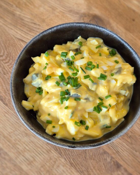 Cremiger Eiersalat mit gehackten Eiern, Gurkenwürfeln und feinen Schnittlauchstreifen in einer schwarzen Schüssel auf einem Holztisch.