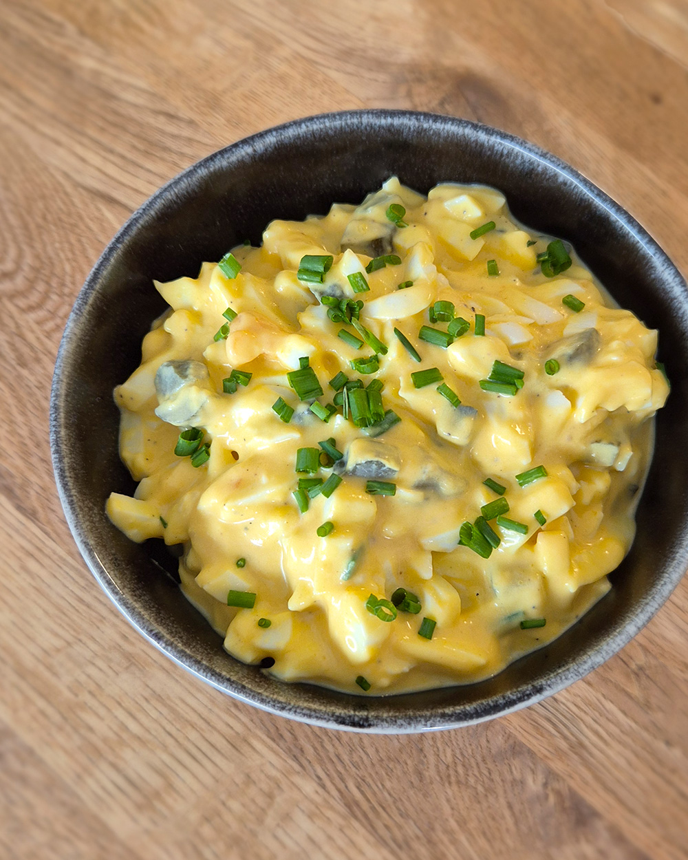 Cremiger Eiersalat mit gehackten Eiern, Gurkenwürfeln und feinen Schnittlauchstreifen in einer schwarzen Schüssel auf einem Holztisch.