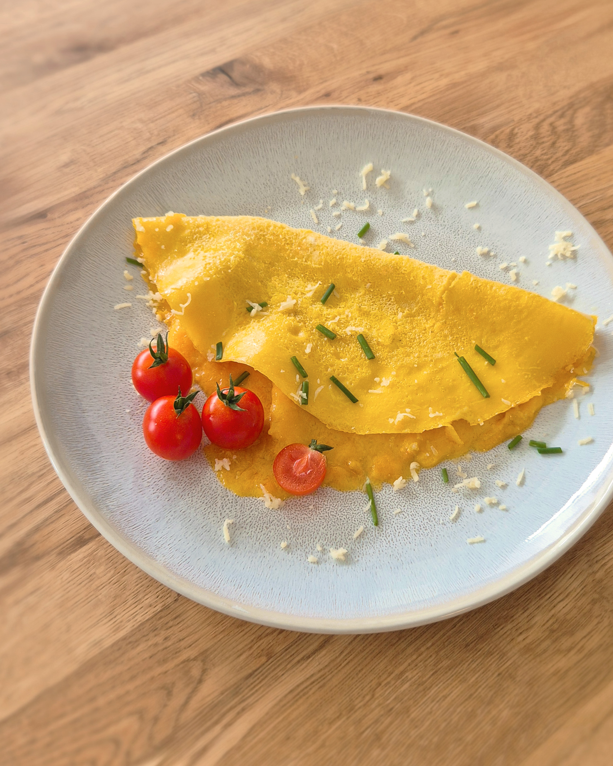 Käse-Omelette auf einem hellen Teller, garniert mit Kirschtomaten und feinem Schnittlauch, Käseflocken darüber, Holzuntergrund, appetitliches Frühstücksbild aus Bettys Küchenschätze