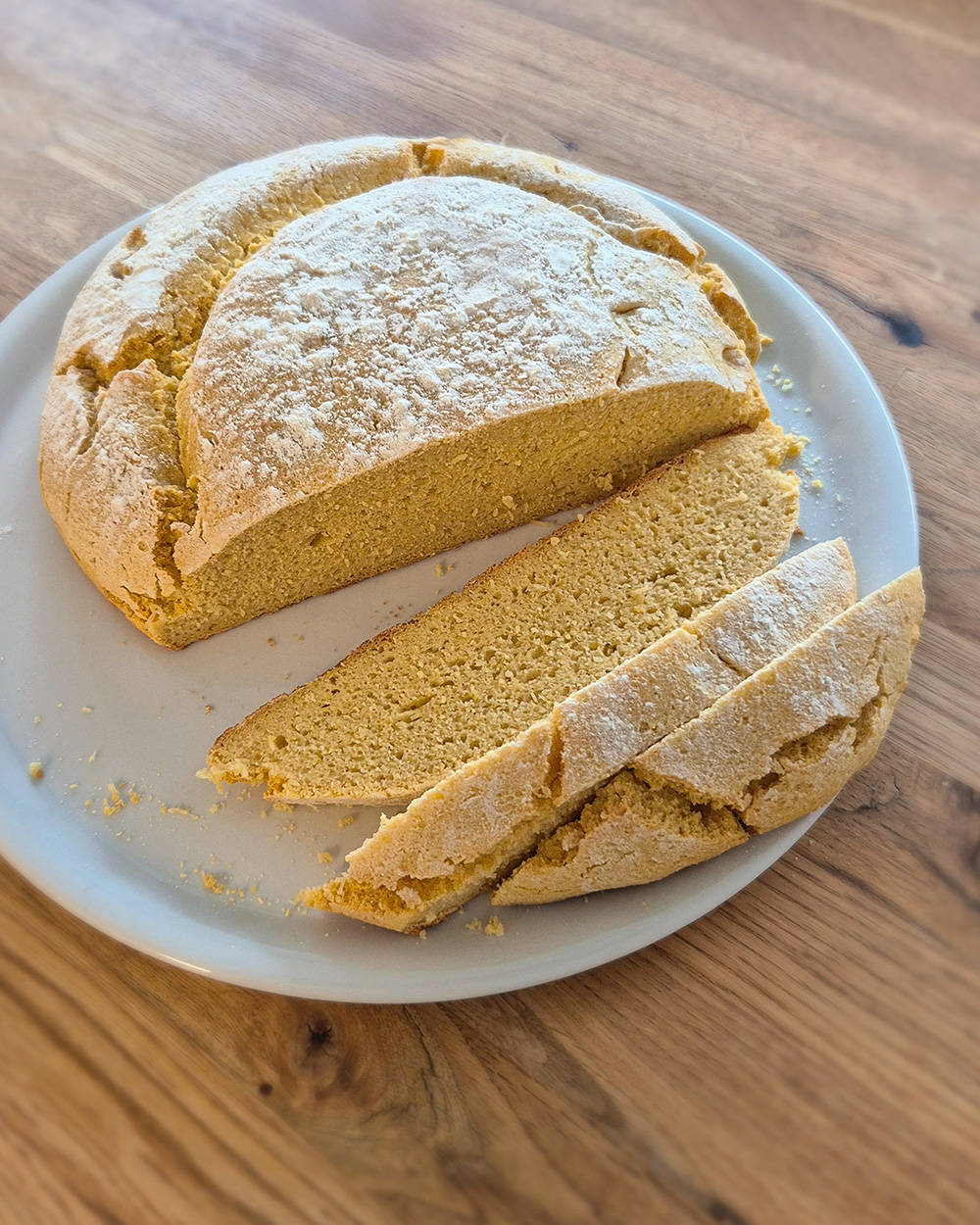 Portugiesisches Maisbrot mit goldgelber Kruste, leicht mehlbestäubt, davon drei Scheiben abgeschnitten, auf dem weißen Teller, Rezeptbild aus Betty's Küchenschätze.