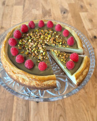 Pistazien-Cheesecake mit pistaziengrüner Creme, knusprigem Keksboden, garniert mit vielen Himbeeren und gehackten Pistazien.