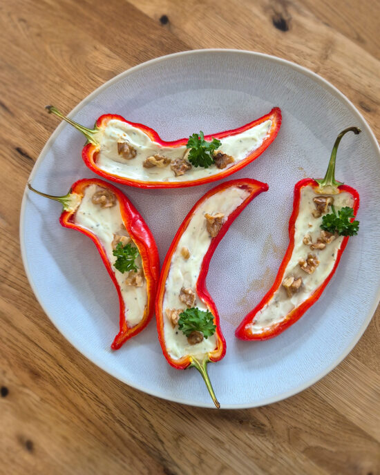 Auf einem hellen Teller liegen gefüllte rote Spitzpaprika mit cremiger Kräuterfrischkäse- und Magerquarkfüllung, bestreut mit Walnüssen und Petersilie.