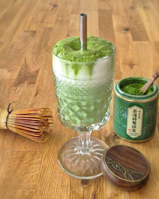 Mit grünem Matcha-Schaum, Milch und Eiswürfeln im dekorativen Glas präsentiert sich der Iced Matcha Latte als kühler Sommerdrink, daneben Bambusbesen und Grünteepulver.
