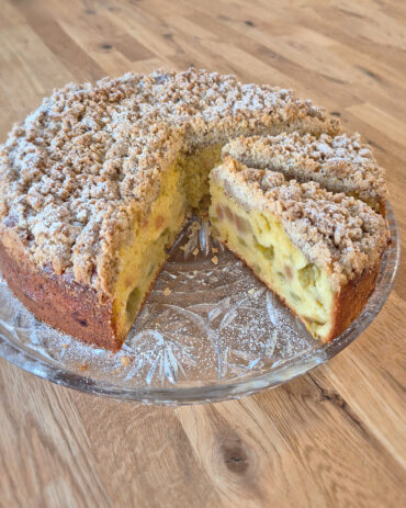 Goldbrauner Rhabarber-Streuselkuchen mit knusprigen Mandelstreuseln, saftigem Teig und fruchtig-säuerlicher Rhabarber auf einer Tortenplatte.