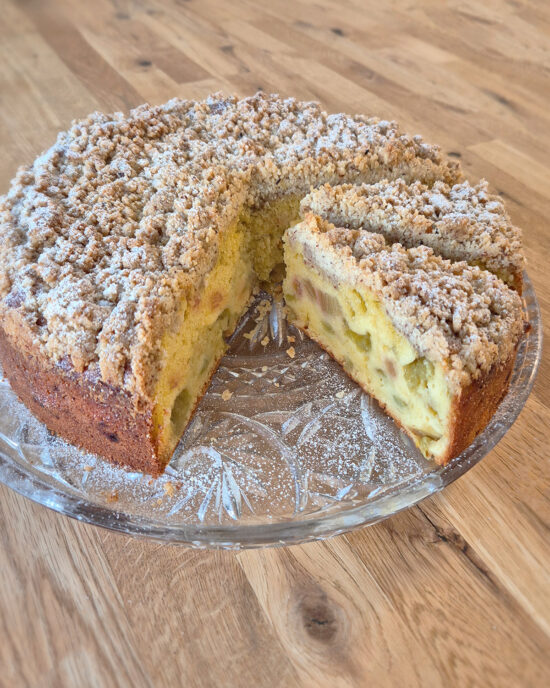 Goldbrauner Rhabarber-Streuselkuchen mit knusprigen Mandelstreuseln, saftigem Teig und fruchtig-säuerlicher Rhabarber auf einer Tortenplatte.