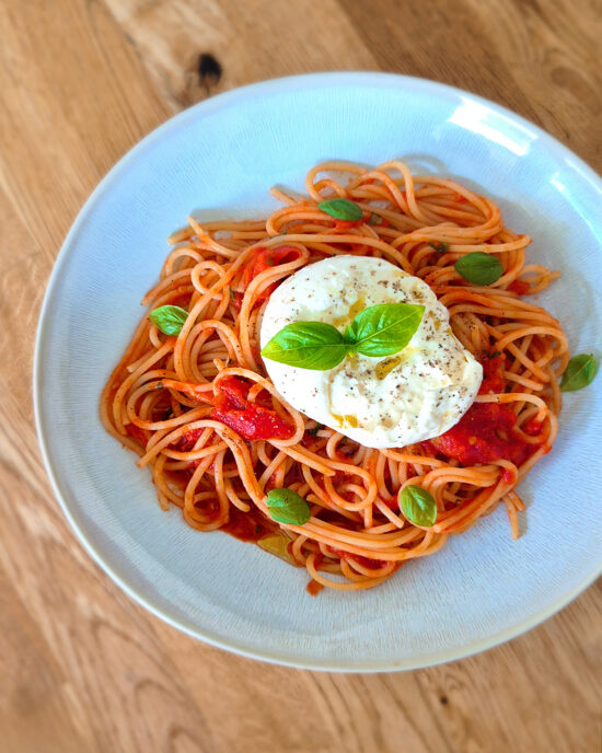 Auf dem Teller liegen Tomatenspaghetti mit würziger Tomatensauce, frischem Basilikum und cremiger Burrata, leicht mit Pfeffer und Öl verfeinert.