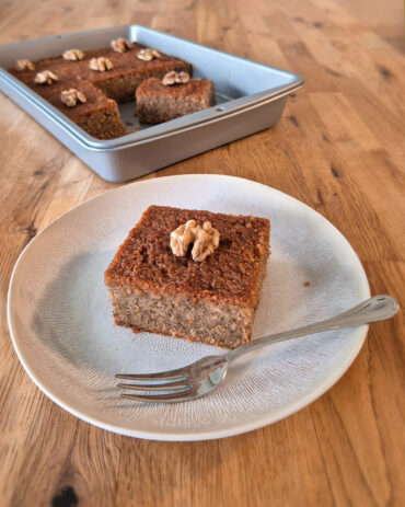 Saftiger Karydopita, griechischer Walnusskuchen mit Honigsirup, Zimt und Walnüssen, in einer rechteckigen Backform und in quadratisches Stück auf einem Teller angerichtet.