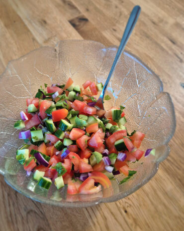 In einer Glasschale liegt frischer Kachumber Salat mit gewürfelten Gurken, Tomaten, roten Zwiebeln und Koriander auf einem Holztisch.