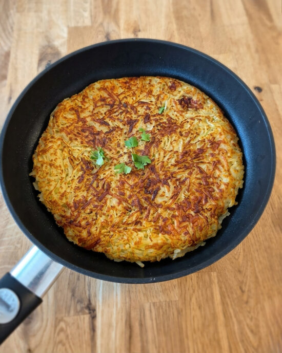Goldbraune Schweizer Rösti mit knuspriger Oberfläche liegt in einer schwarzen Pfanne auf Holz, in der Mitte mit frischer Petersilie garniert.