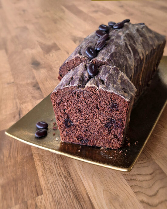 Ein saftiger Schokoladen-Kaffeekuchen mit Espressoglasur und Schokostückchen, als Cake gebacken, auf goldener Platte serviert und ein Stück abgeschnitten