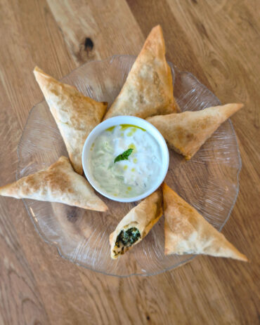 Goldbraune Spanakopita-Dreiecke liegen auf einem Glasteller rund um einen Tzatziki-Dip mit Minze und Olivenöl, auf einem Holztisch.