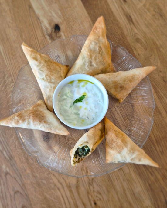 Goldbraune Spanakopita-Dreiecke liegen auf einem Glasteller rund um einen Tzatziki-Dip mit Minze und Olivenöl, auf einem Holztisch.