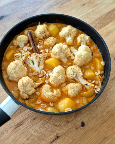 In einer schwarzen Pfanne ist gerösteter Blumenkohl in orangefarbener Massaman-Currysauce mit Kartoffeln, Erdnüssen und einer Zimtstange.