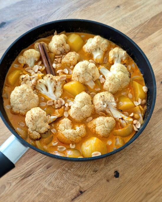 In einer schwarzen Pfanne ist gerösteter Blumenkohl in orangefarbener Massaman-Currysauce mit Kartoffeln, Erdnüssen und einer Zimtstange.