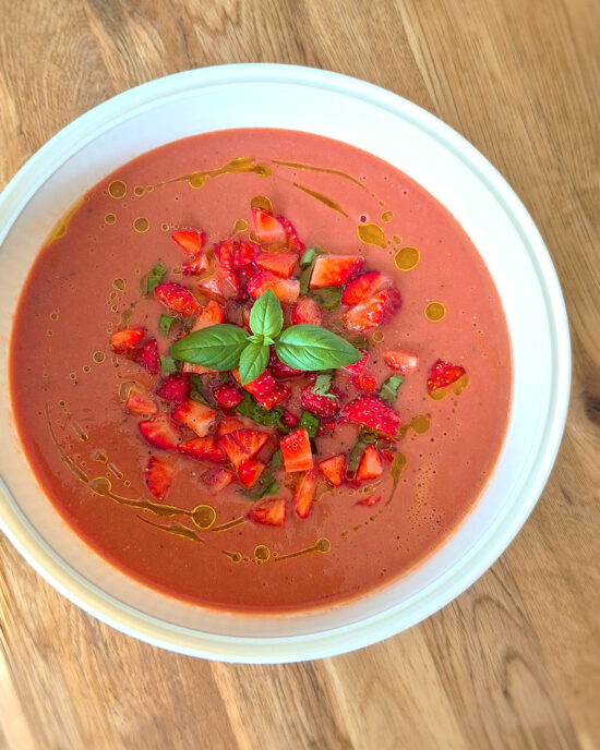 Eine weisse Schale Erdbeer-Gazpacho mit Erdbeerstückchen, Basilikum und Olivenöl steht auf einem Holztisch.