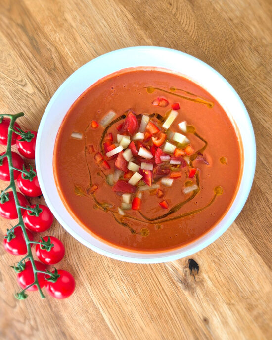 Auf einem Holztisch steht Gazpacho andaluz, getoppt mit Tomatenwürfeln, Gurke, Paprika und Olivenöl, daneben liegen frische Cherrytomaten am Zweig.