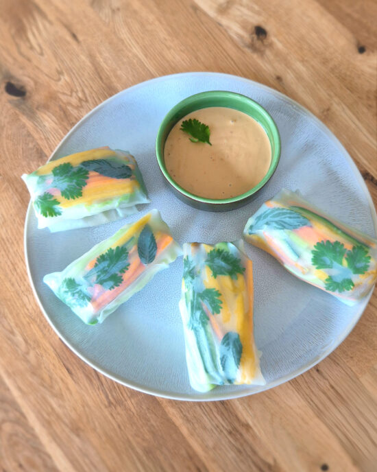 Bunte Hähnchen-Sommerrollen mit Mango, Karotten, Gurken und Kräutern liegen auf einem Teller neben cremigem Erdnussdip.