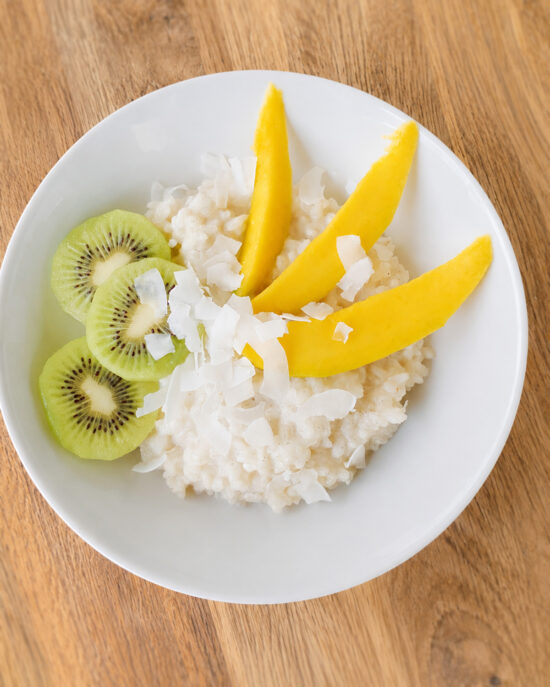 In einer weißen Schale liegt cremiger Kokosmilchreis mit Mango, Kiwi und Kokoschips auf einem Holztisch.