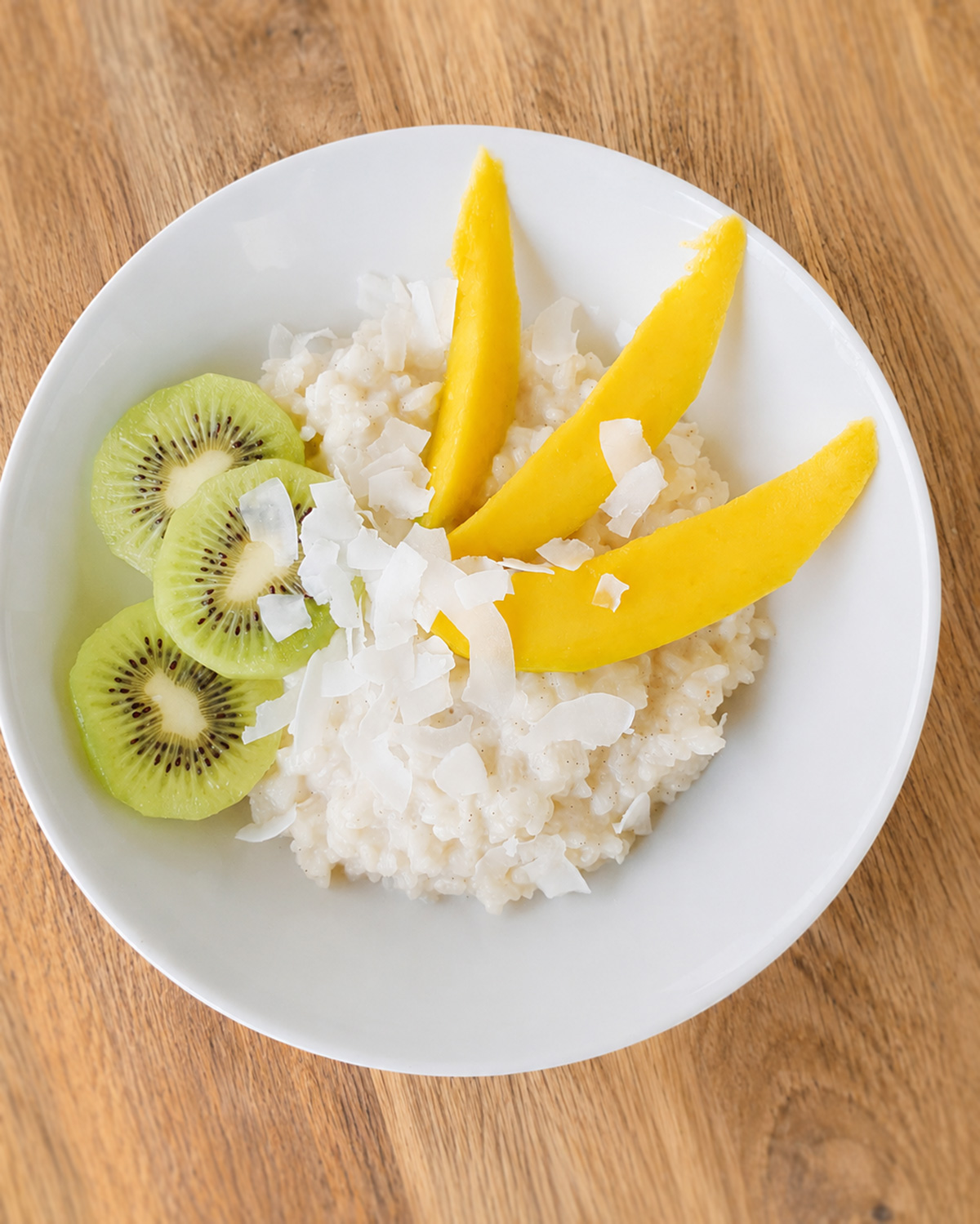 In einer weißen Schale liegt cremiger Kokosmilchreis mit Mango, Kiwi und Kokoschips auf einem Holztisch.