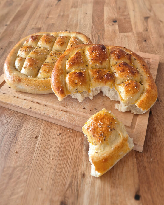 Auf einem Holzbrett liegen zwei goldbraune Pide mit Sesam und Schwarzkümmel, daneben liegt ein abgebrochenes Stück mit weicher, fluffiger Krume.