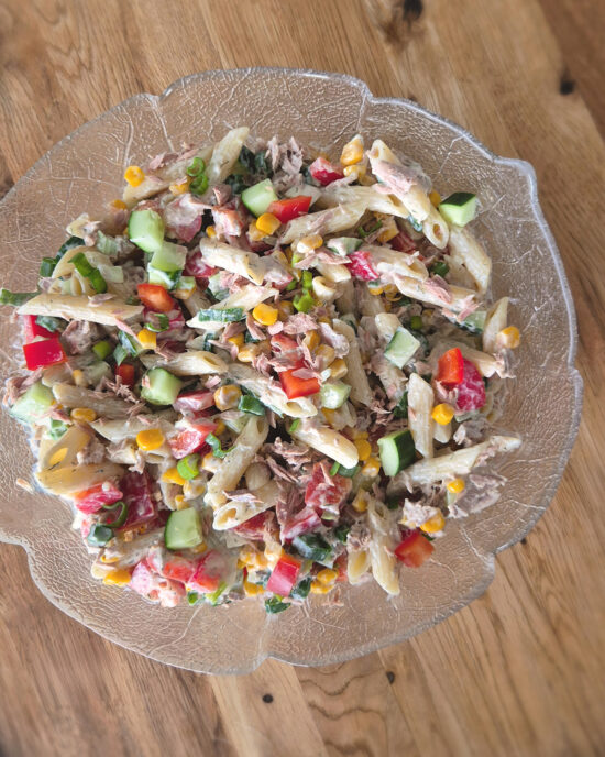 Cremiger Thunfisch-Nudelsalat mit Penne, Mais, Gurke, roter Paprika und Frühlingszwiebeln liegt in einer Glasschale auf einem Holztisch.