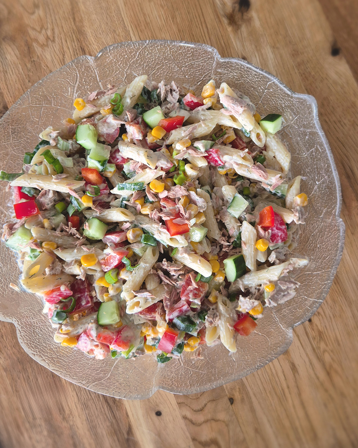 Cremiger Thunfisch-Nudelsalat mit Penne, Mais, Gurke, roter Paprika und Frühlingszwiebeln liegt in einer Glasschale auf einem Holztisch.