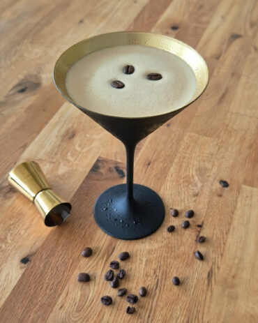 Gekühlter Espresso Martini mit cremiger Schaumkrone, drei Kaffeebohnen und verstreuten Bohnen, serviert in einem schwarz-goldenen Martiniglas auf einem Holztisch