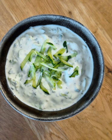 Cremiges Gurken-Raita mit frischen Gurkenstreifen und Kräutern in einer schwarzen Schale auf einem Holztisch