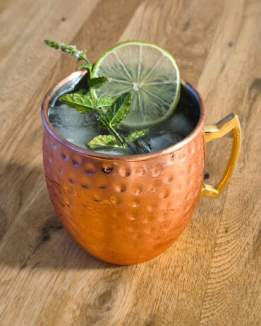Erfrischender London Mule mit Eiswürfeln, frischer Minze und Limettenscheibe im kupfernen Becher auf einem Holztisch