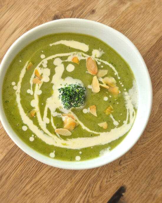 Cremige Brokkolisuppe mit Rahm, Mandelblättchen und einem kleinen Brokkoliröschen in einer weißen Schale auf einem Holztisch.