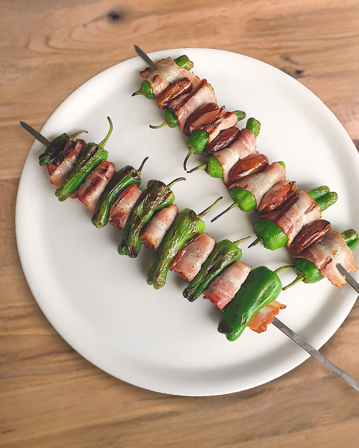 Auf einem weissen Teller liegen gegrillte Pimientos-Spiesse mit süssen Datteln und würzigem Speck, der die Zutaten umwickelt.